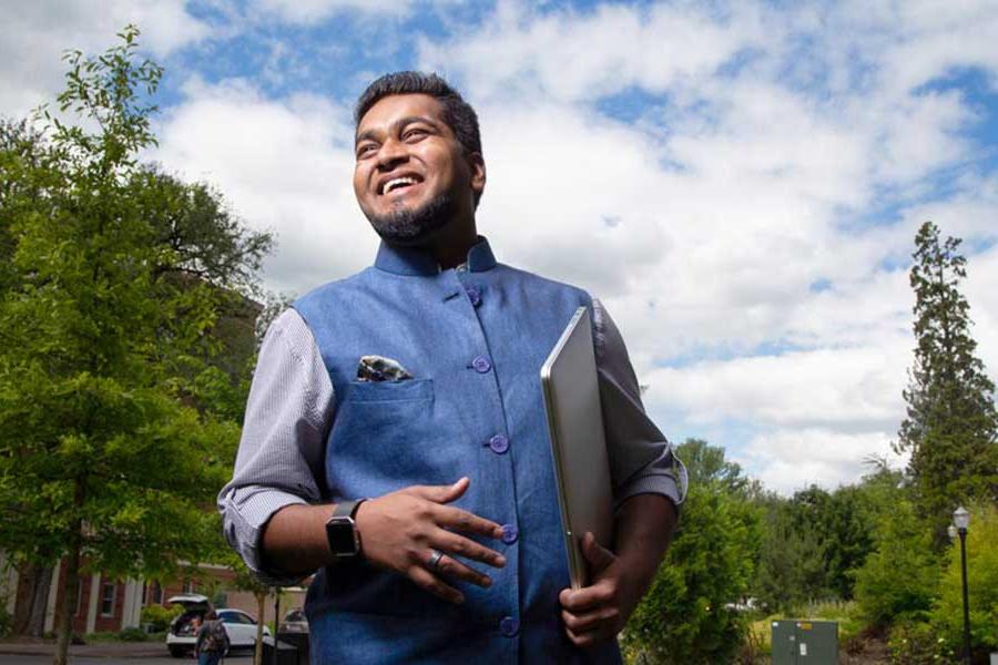 person holding a laptop wearing a blue vest and shirt standing in front of fir trees